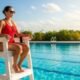 A lifeguard, wearing a red swimsuit and a whistle, sits attentively on a high chair overlooking a sparkling blue swimming pool on a sunny day. They are focused on the water, ready to act.